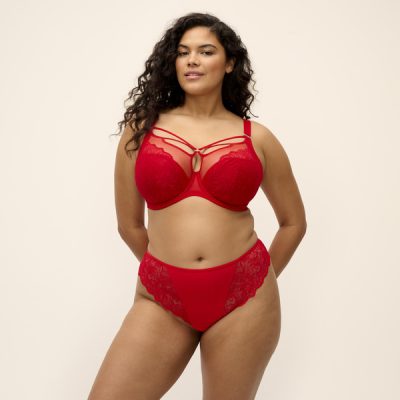 BRIANNA_HAUTE-RED_UW-PLUNGE-BRA_EL8080_THONG_EL8085_F_TRADE_3000_SS27 Kobieta o pełnych kształtach w czerwonym komplecie: biustonosz z fiszbinami z paseczkami na dekolcie i koronkowe figi